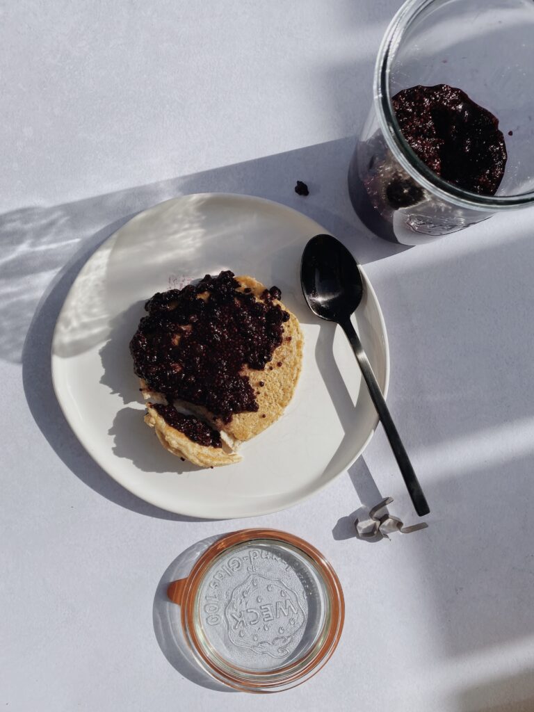 Pancake topped with dark purple blueberry chia jam on a white plate with a black spoon. In the upper right-hand corner is a glass jar of opened blueberry chia jam.
