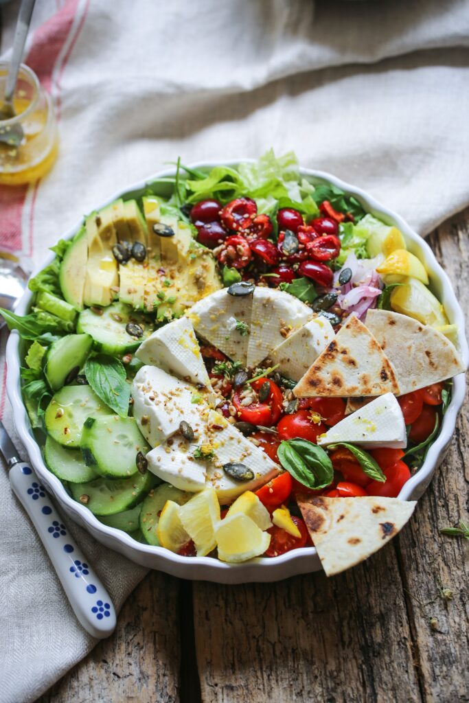 Big salad with leafy greens, cucumber, tomatoes, olives, artichokes, sliced avocado, pumpkin seeds, a wheel of cheese, pita, lemon, onion, basil, and olive oil.