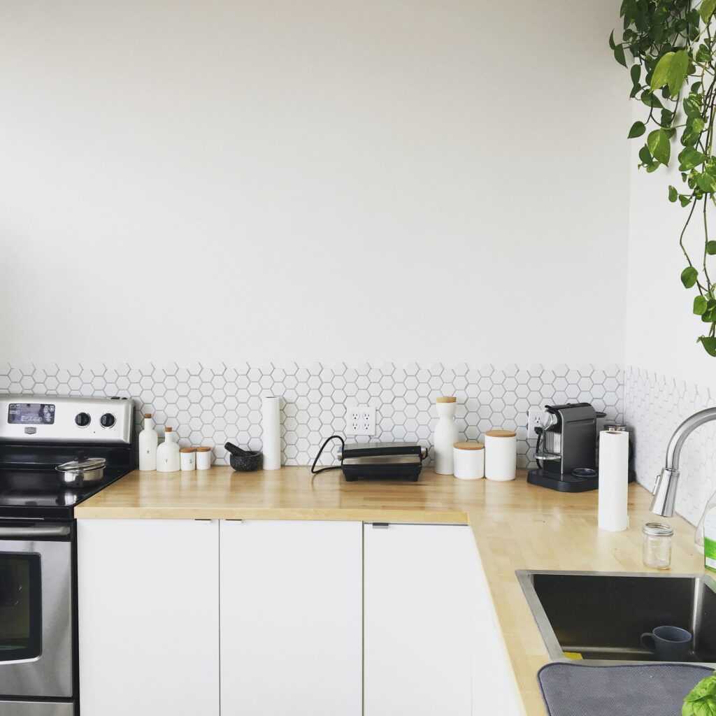 Simple, modern kitchen with a white backsplash and wood countertops. On the counter are white containers for herbs and spices, a small coffee maker, a stainless steel sink, and a roll of paper towels. A green plant hangs from an upper shelf.